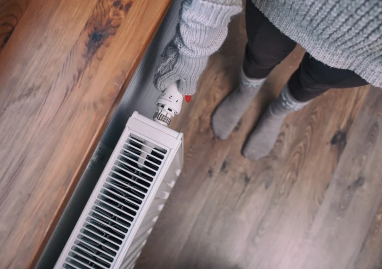 Person wearing gloves and socks adjusts a radiator valve in a room with wooden flooring.