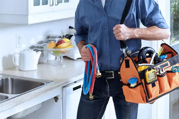 Person in blue shirt holding a plumbing tool bag and hoses in a kitchen near a sink.